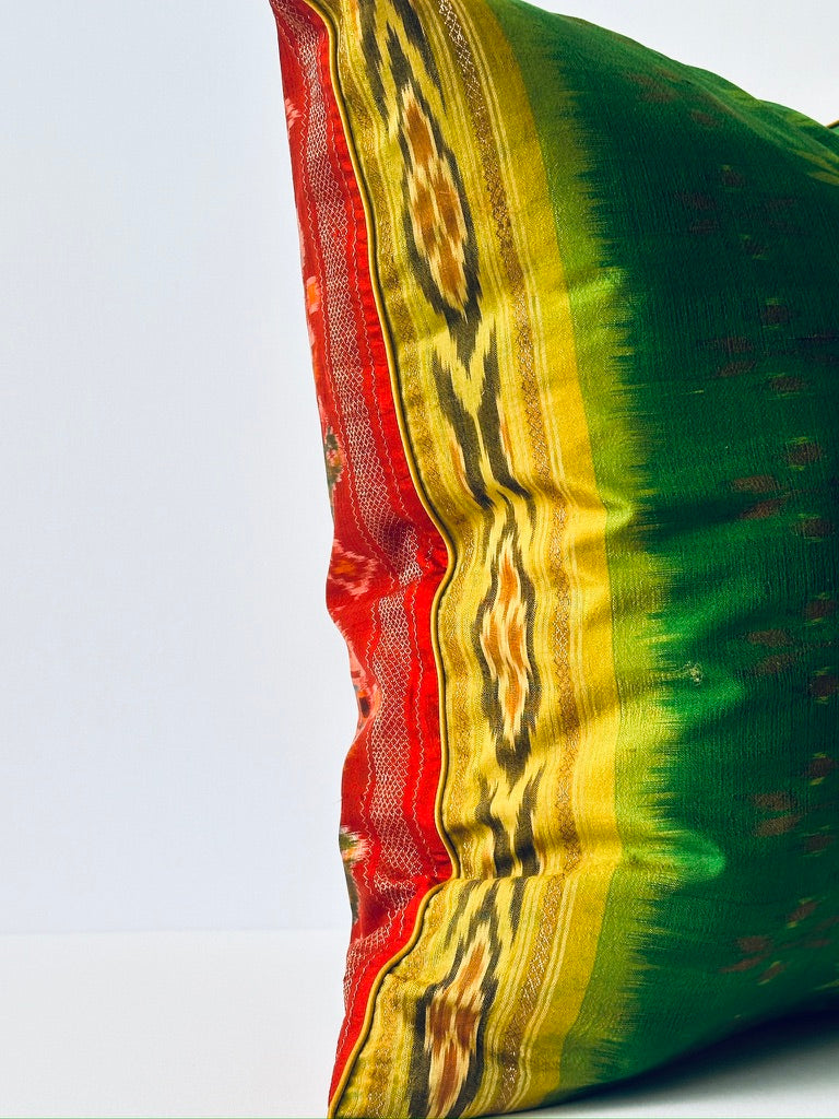 A handcrafted pillow cover made from traditional fabric with colorful motifs in green, red, and yellow on a cream background. The cushion is adorned with a bold red border featuring intricate geometric patterns, adding a vibrant and cultural touch to any room.