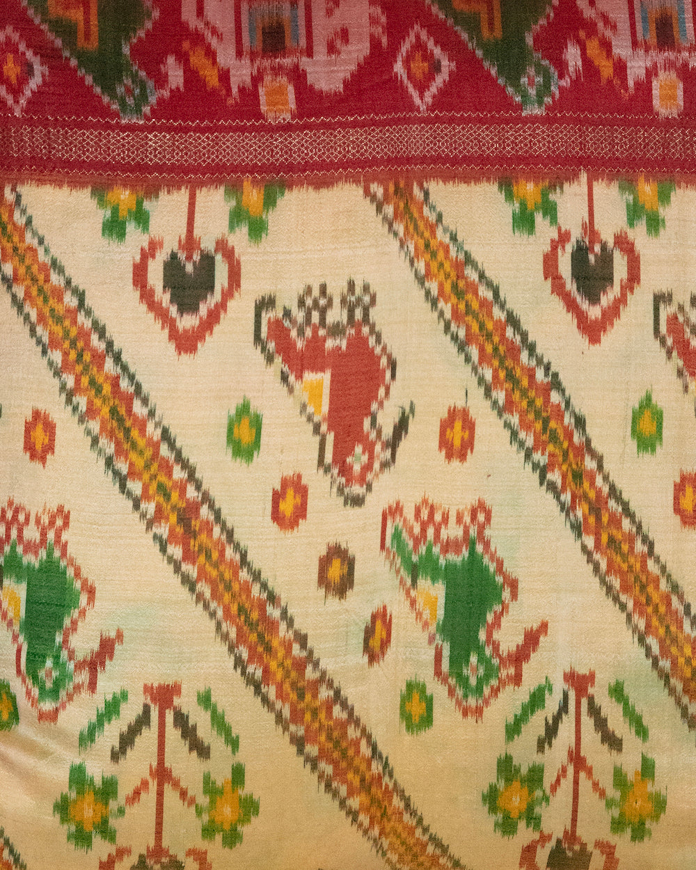 A handcrafted pillow cover made from traditional fabric with colorful motifs in green, red, and yellow on a cream background. The cushion is adorned with a bold red border featuring intricate geometric patterns, adding a vibrant and cultural touch to any room.