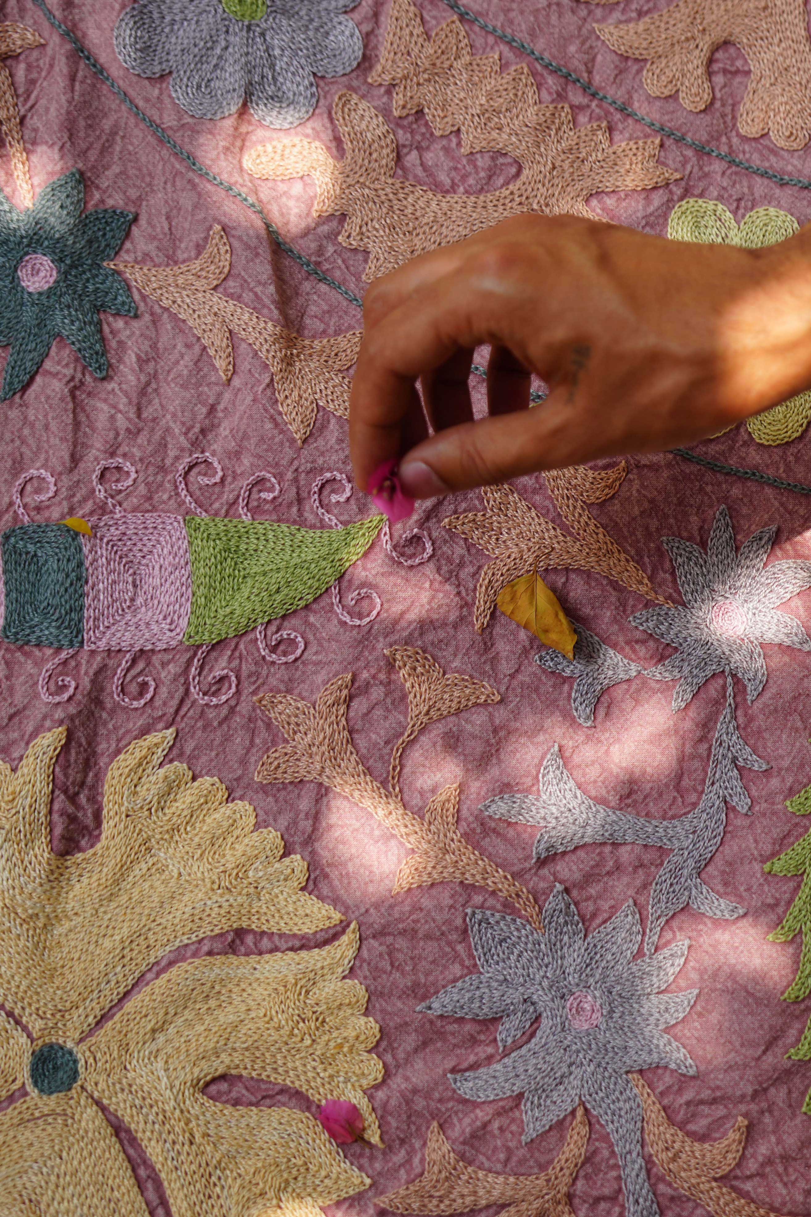 A detailed view of a vintage Suzani textile draped elegantly, featuring intricate hand-embroidered floral and geometric designs in soft pink, yellow, green, and blue hues. The fabric showcases traditional craftsmanship with its rich texture and vibrant colors, adding a touch of cultural elegance.