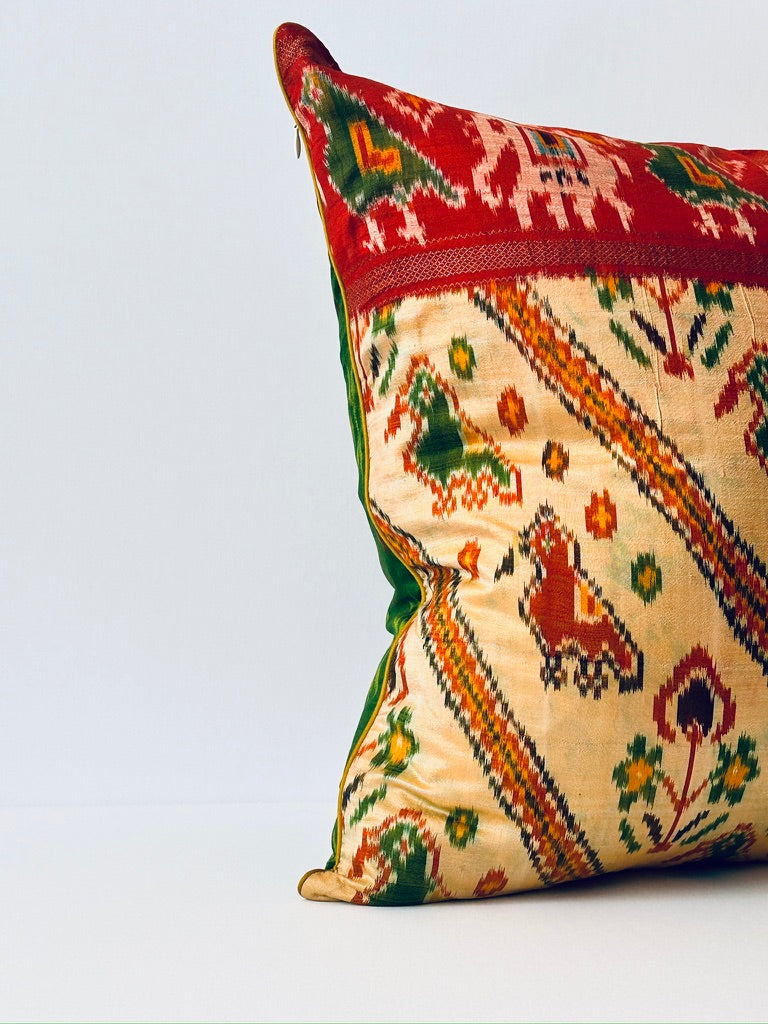 A handcrafted pillow cover made from traditional fabric with colorful motifs in green, red, and yellow on a cream background. The cushion is adorned with a bold red border featuring intricate geometric patterns, adding a vibrant and cultural touch to any room.