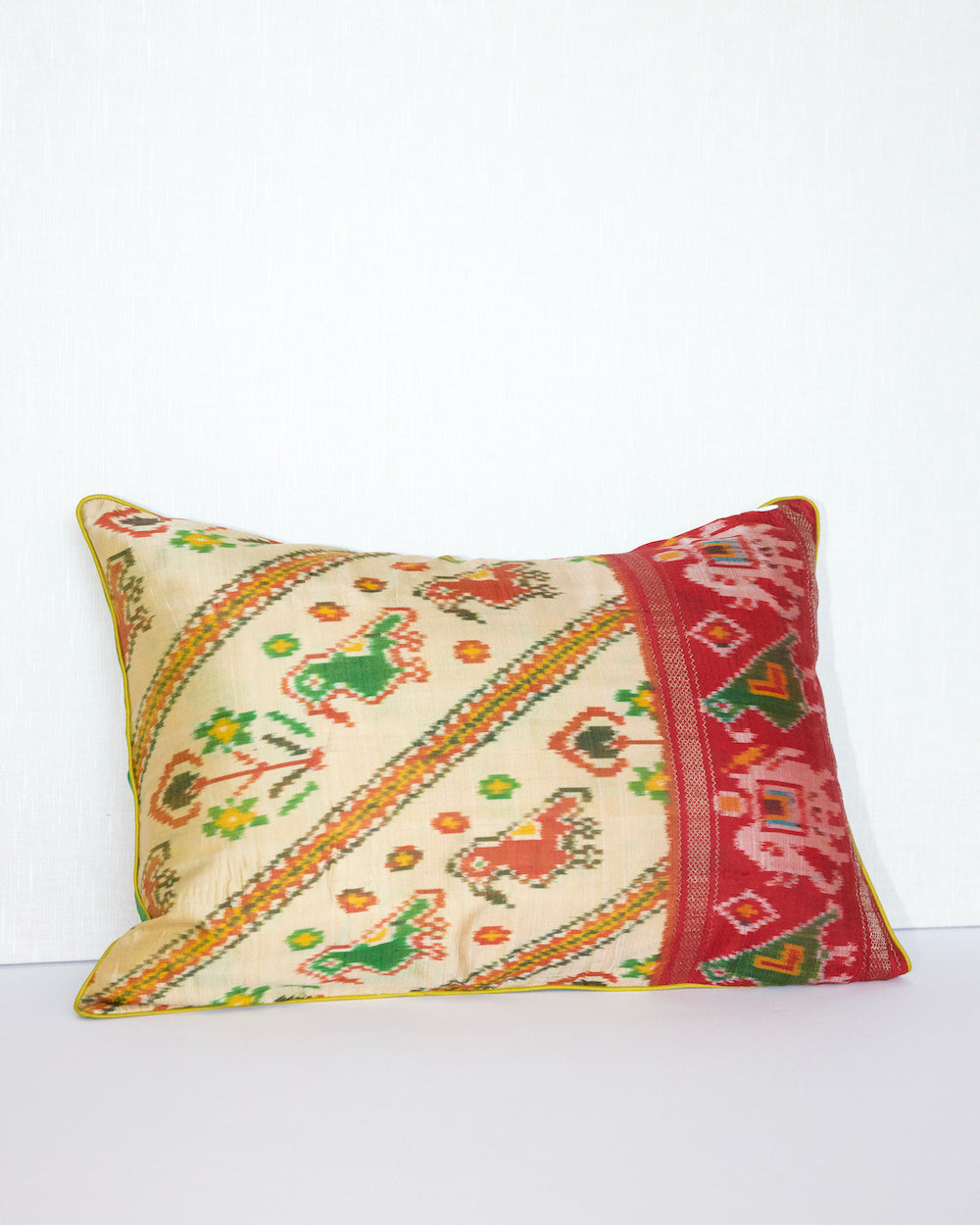 A handcrafted pillow cover made from traditional fabric with colorful motifs in green, red, and yellow on a cream background. The cushion is adorned with a bold red border featuring intricate geometric patterns, adding a vibrant and cultural touch to any room.