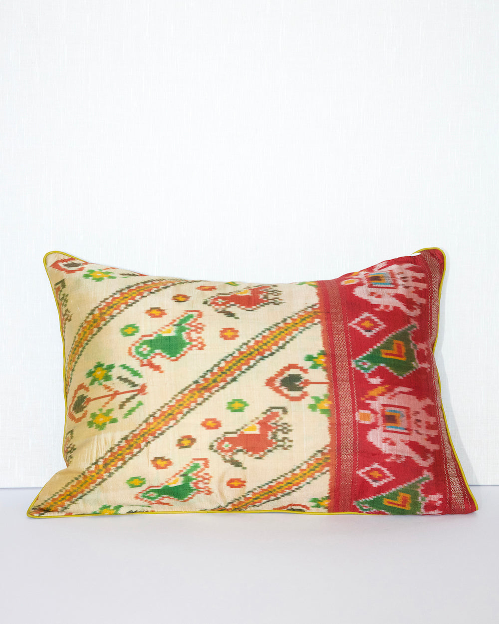 A handcrafted pillow cover made from traditional fabric with colorful motifs in green, red, and yellow on a cream background. The cushion is adorned with a bold red border featuring intricate geometric patterns, adding a vibrant and cultural touch to any room.