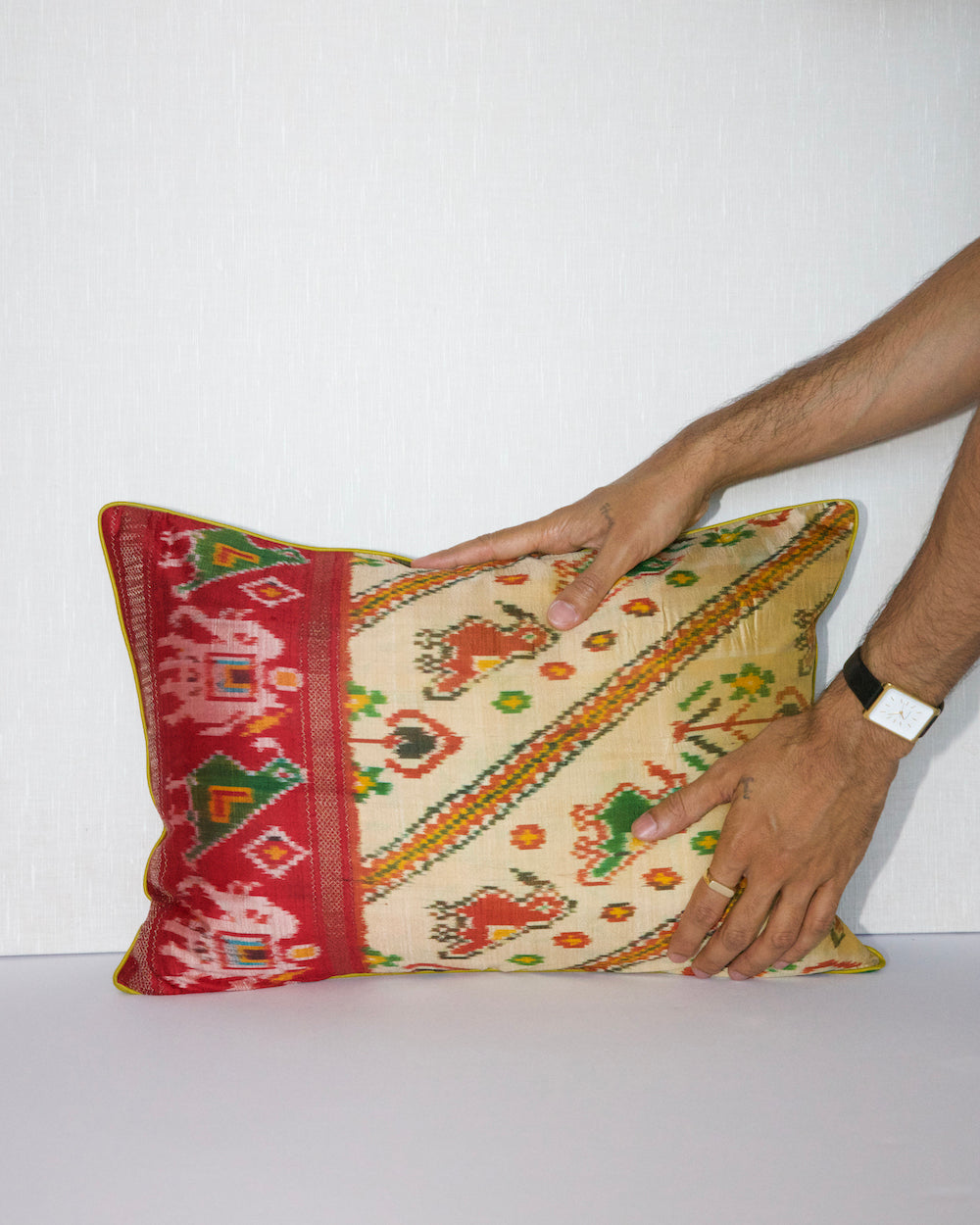 A handcrafted pillow cover made from traditional fabric with colorful motifs in green, red, and yellow on a cream background. The cushion is adorned with a bold red border featuring intricate geometric patterns, adding a vibrant and cultural touch to any room.