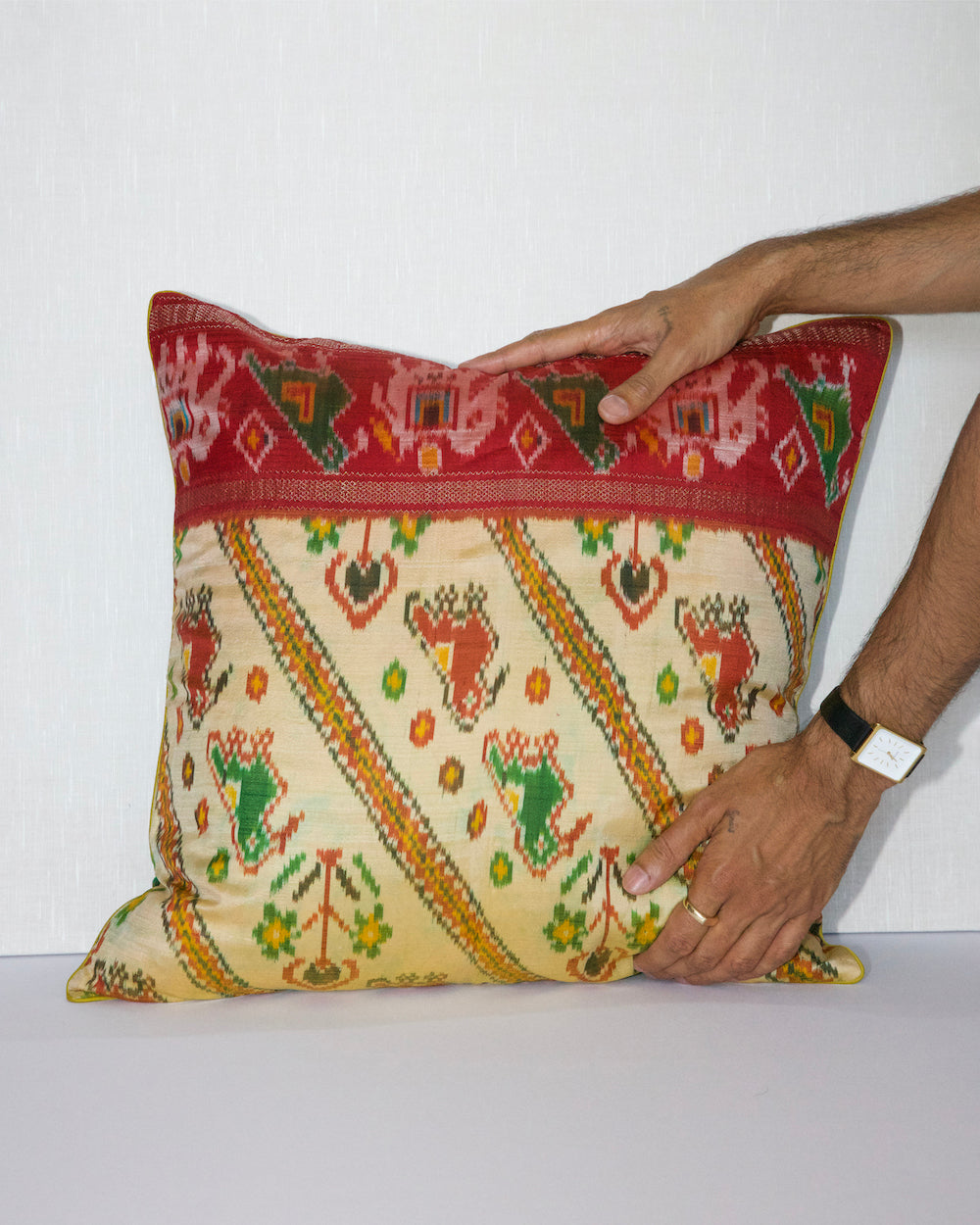 A handcrafted pillow cover made from traditional fabric with colorful motifs in green, red, and yellow on a cream background. The cushion is adorned with a bold red border featuring intricate geometric patterns, adding a vibrant and cultural touch to any room.