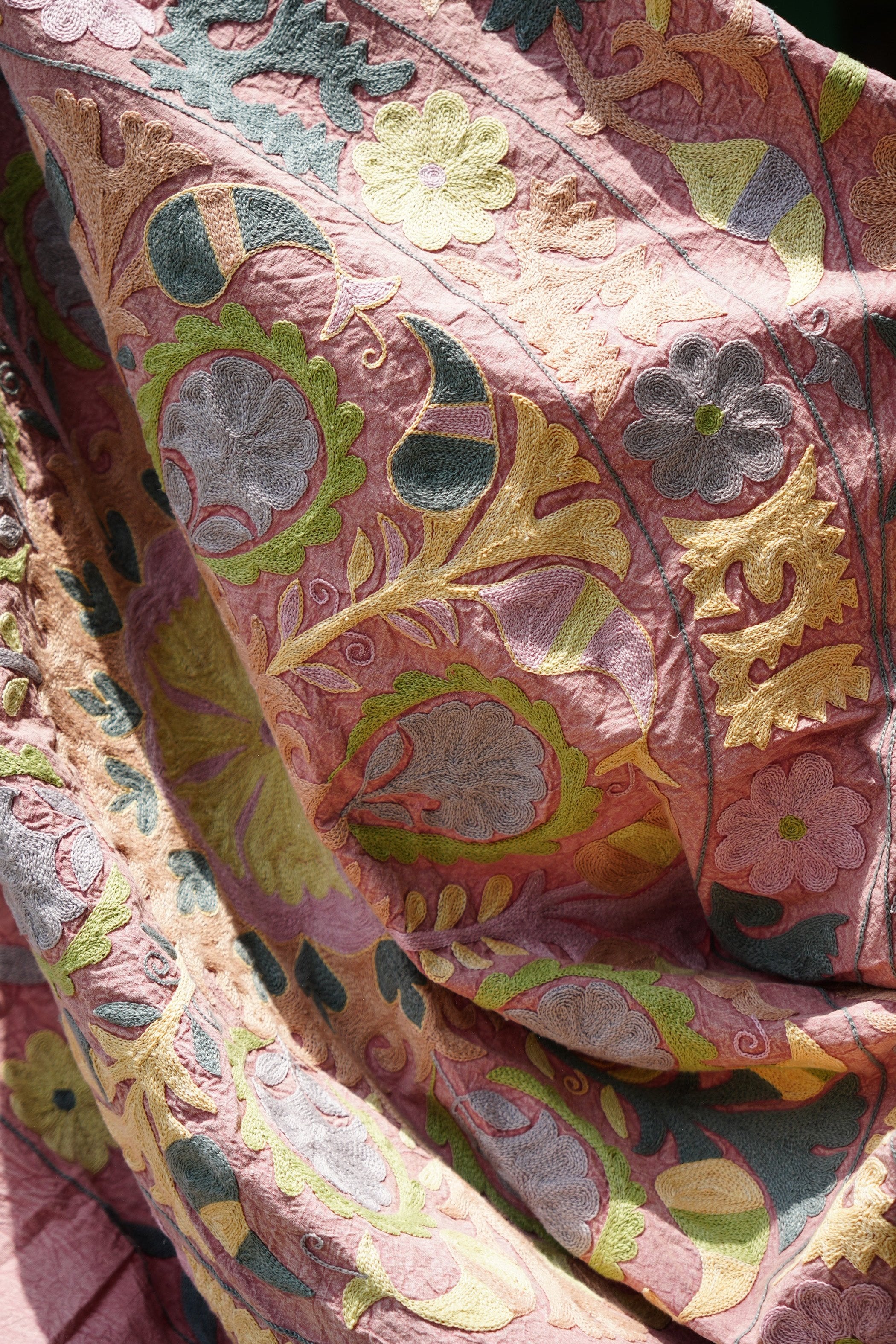 A detailed view of a vintage Suzani textile draped elegantly, featuring intricate hand-embroidered floral and geometric designs in soft pink, yellow, green, and blue hues. The fabric showcases traditional craftsmanship with its rich texture and vibrant colors, adding a touch of cultural elegance.