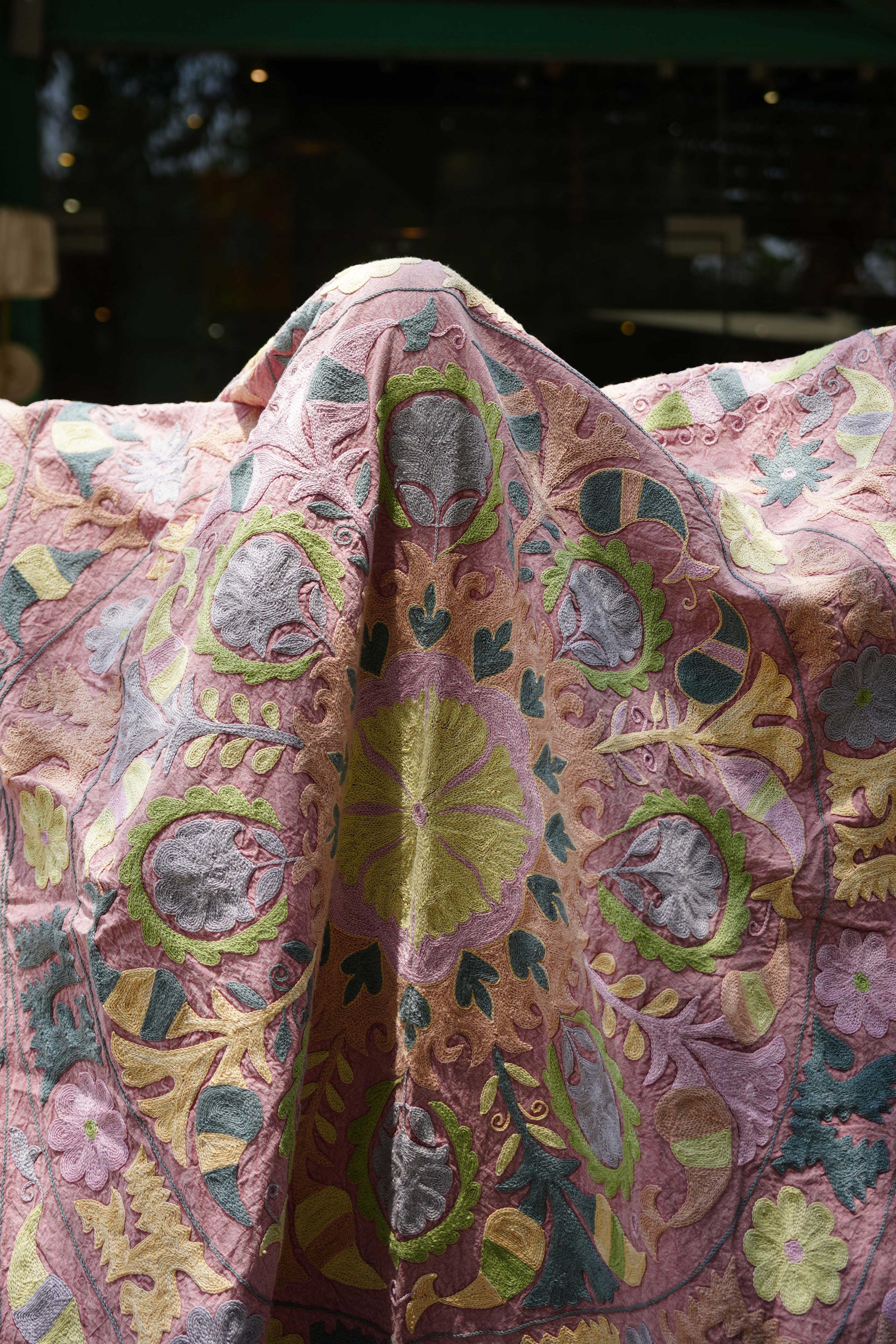 A detailed view of a vintage Suzani textile draped elegantly, featuring intricate hand-embroidered floral and geometric designs in soft pink, yellow, green, and blue hues. The fabric showcases traditional craftsmanship with its rich texture and vibrant colors, adding a touch of cultural elegance.